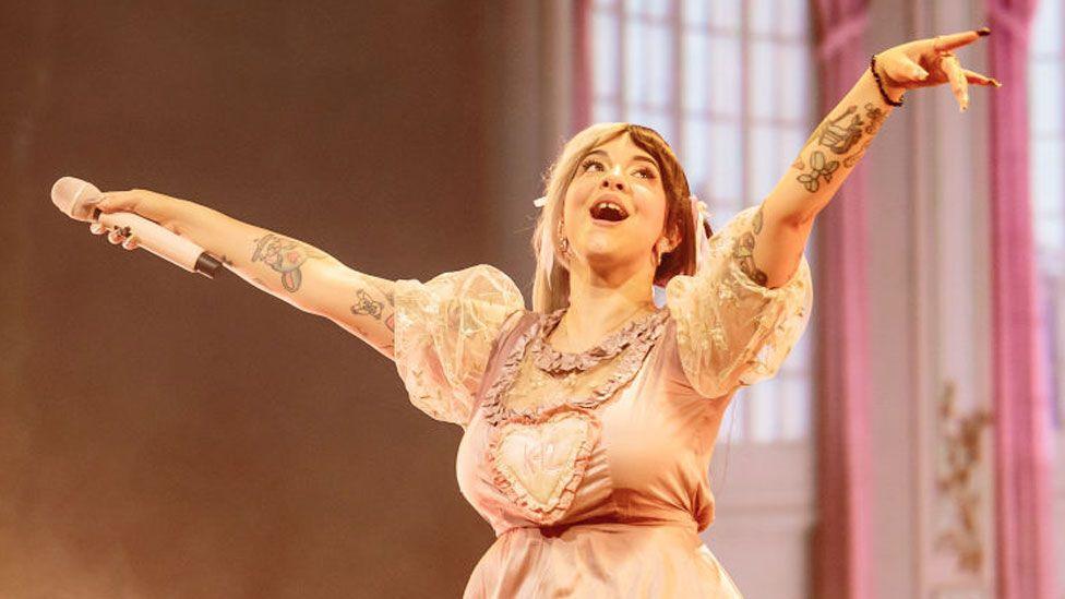 Melanie Martinez perform in 2024 in Milan, Italy. She has her arms outstretched with her microphone held in her right hand and is looking up and smiling. She is wearing a pink dress, with short white puffed sleeves