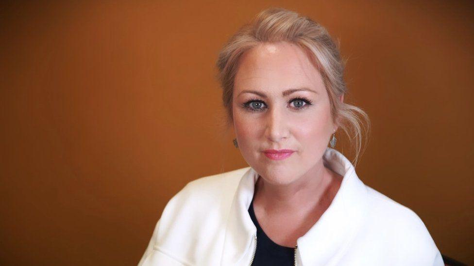 Llinos Griffin-Williams standing in front of an orange background. She is looking at the camera. She has her hair tied back and is wearing a white jacket. 