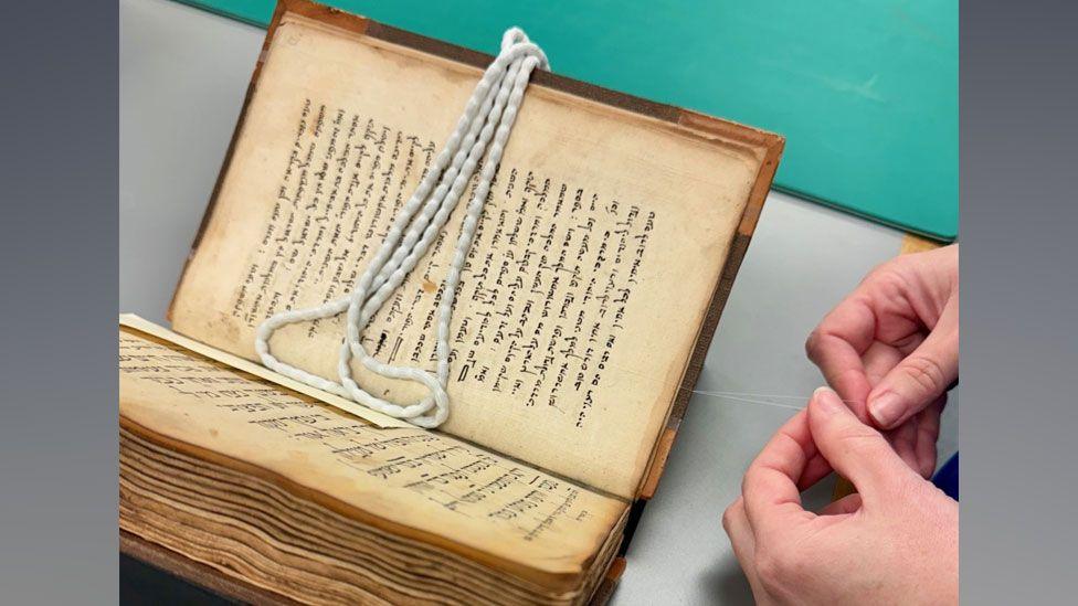 An book showing sepia-coloured pages, showing a Hebrew commentary on the Bible, with two hands holding thread to the right 