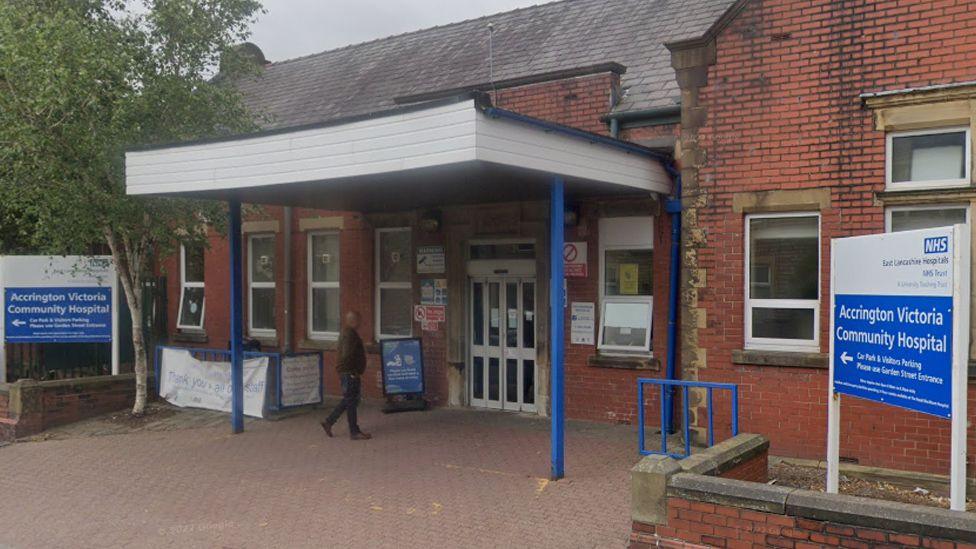 A street view image of Accrington Victoria Hospital, a red brick building with blue and white signs either side of a white awning with blue legs over the entrance and a man walking under it to the front door