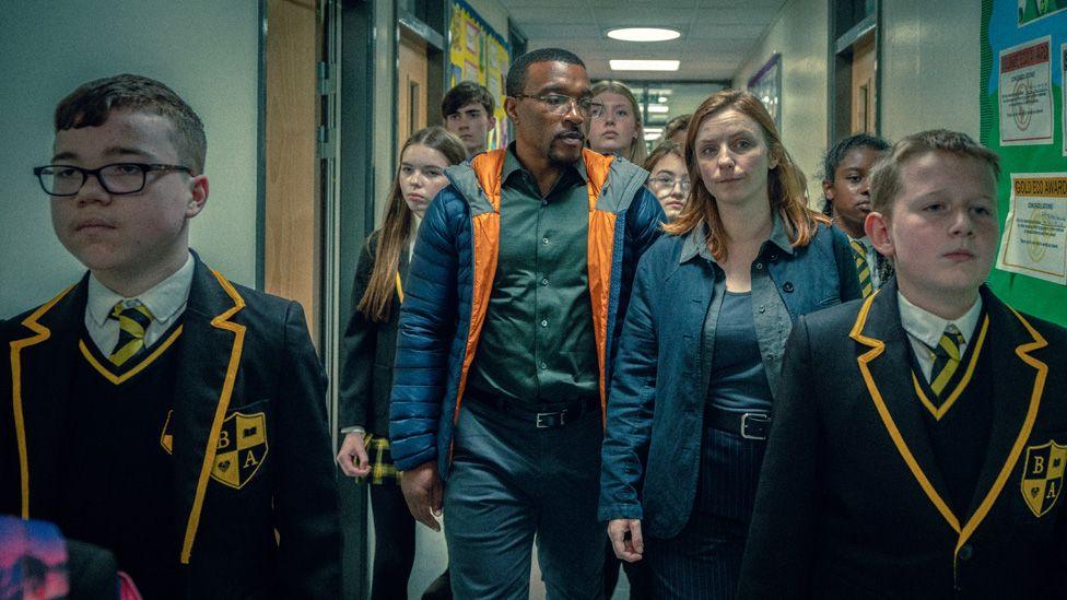 Ashley Walters and Faye Marsay walking down a school corridor at the centre of a group of children in school blazers