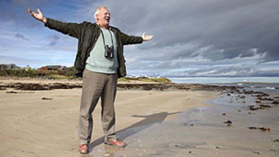 Sir Thomas Allen stands on a beach with his arms outstretched as if singing to the sea