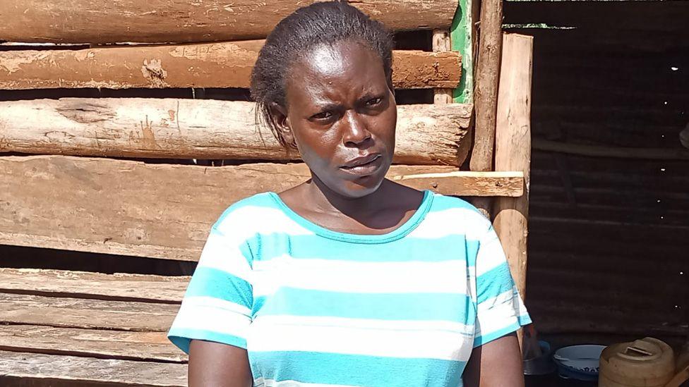 Carolyne Adhiambo in a blue and white horizontal striped T-shirt standing outside a wooden plank house.