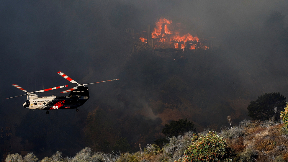  helicopter flies near a burning house as the Palisades Fire rages on at the Mandeville Canyon, in Los Angeles