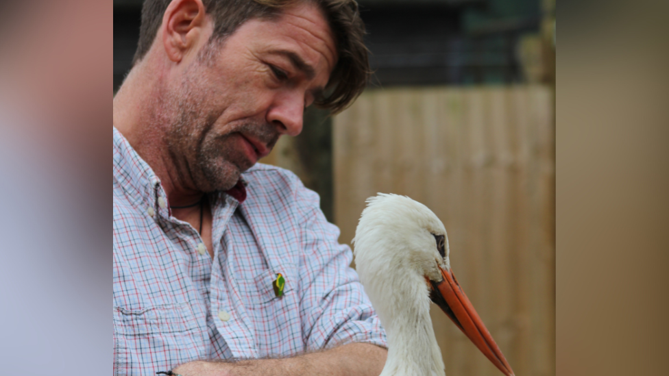 Jamie Craig has short brown hair and brown stubble. He is holding a white stork in his hands.