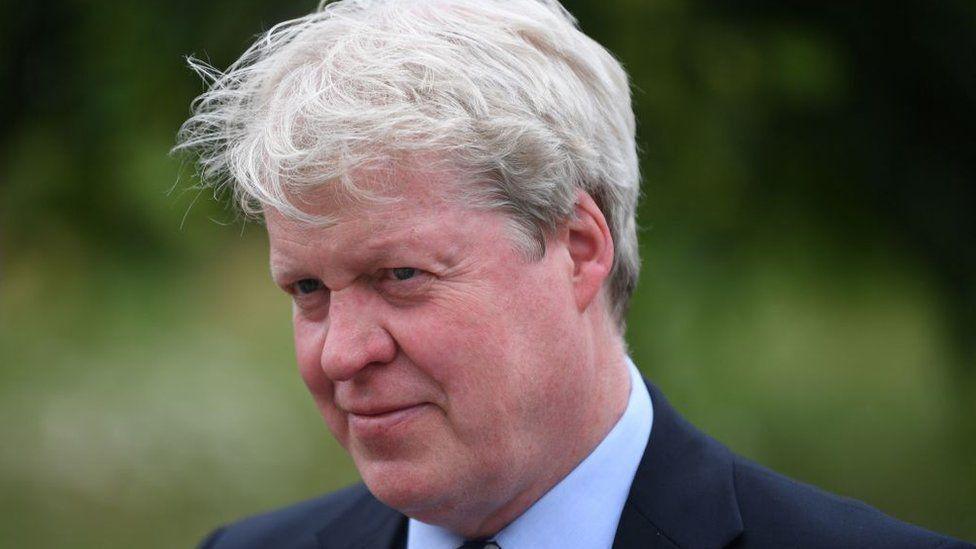 Earl Spencer with greying hair and wearing a dark blue blazer with pale blue  shirt and tie. He is outdoors and and there is greenery behind him.