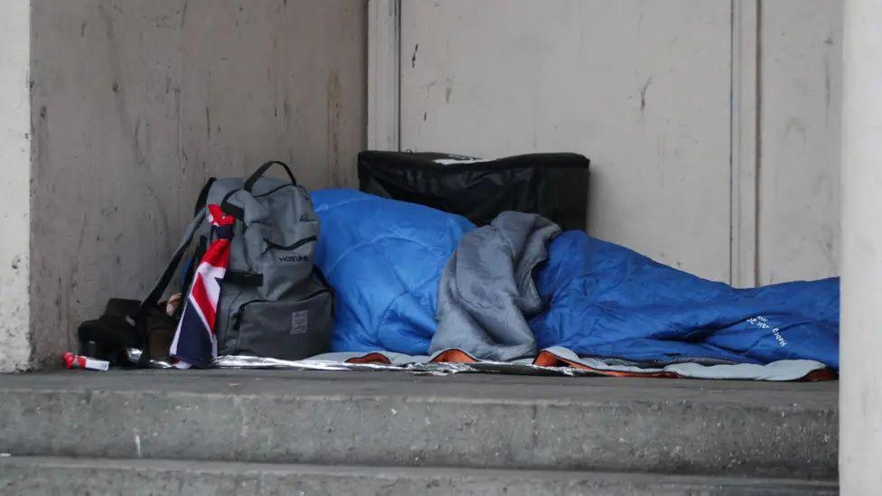 An unidentifiable person wrapped up in a blue sleeping bag on a concrete surface and with a rucksack with Union Flag attached sitting on the ground by their head.