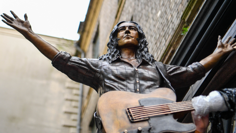 Bronze statue of Rory Gallagher with a guitar hanging off him, photographed against brick wall at Ulster Hall