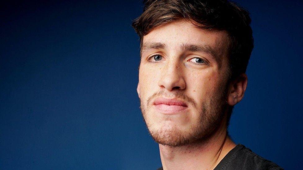 A young man with short dark hair and stubble looks directly at the camera in front of a dark blue background.