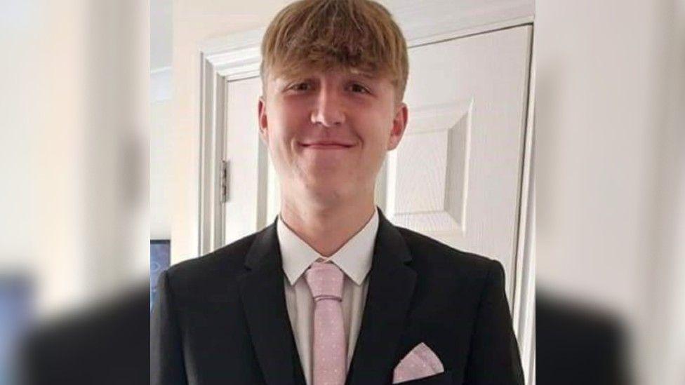 A headshot of Alfie smiling to cameral wearing a dark suit and pink tie