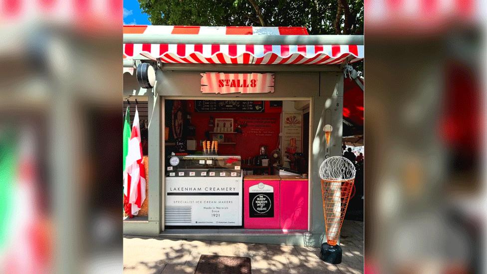A market stall with a red and white striped awning and the sign Stall 8 above a serving hatch. An ice cream fridge has the words Lakenham Creamery written across it. A model ice cream and cone, made of orange wire and white wire, stands about four feet high next to the stall.