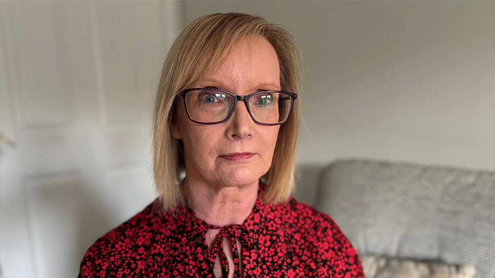 A woman with shoulder length straight blonde hair looks at the camera. She's wearing a black blouse with red flowers