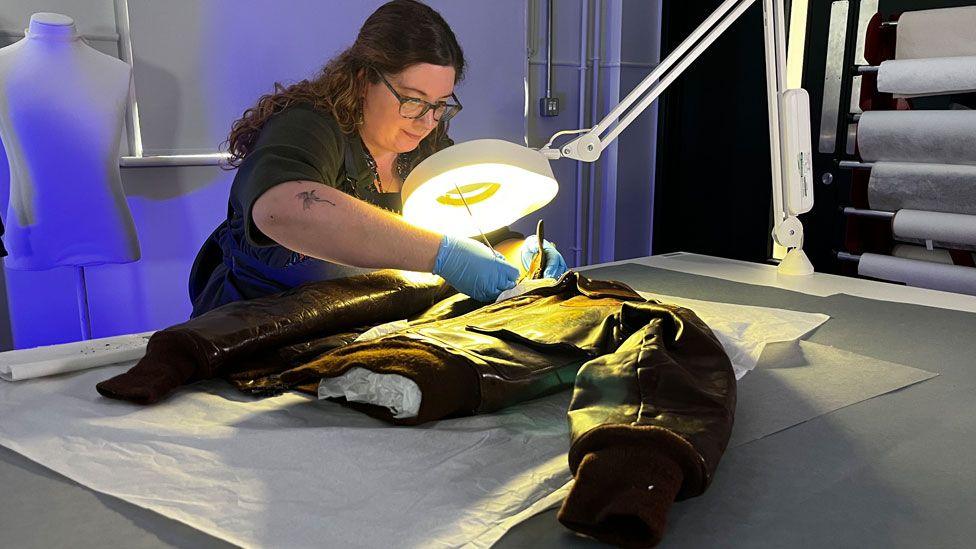 A conservator working an airman's flying jacket. She has long curling dark brown hair pulled back from her face and is wearing glasses, a black T-shirt and blue plastic gloves. She is bending over a table where the jacket is lying on its back, with a bright light shining down upon it. It is resting on tissue paper.