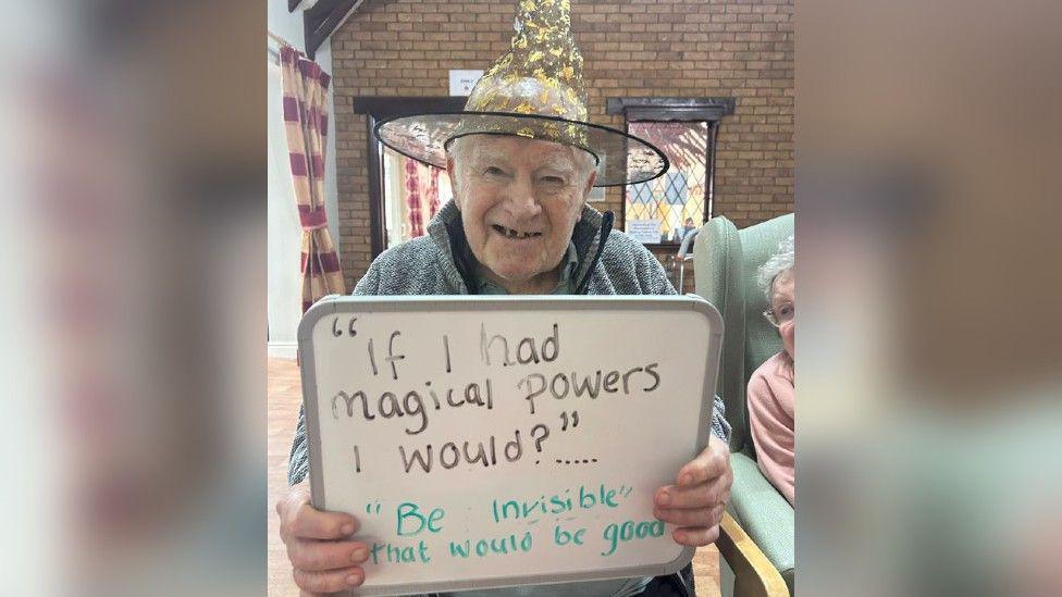 An elderly man sitting down, smiling and wearing a witches hat holding up a sign that says "if I had magical powers I would?" with the answer "be invisible, that would be good".