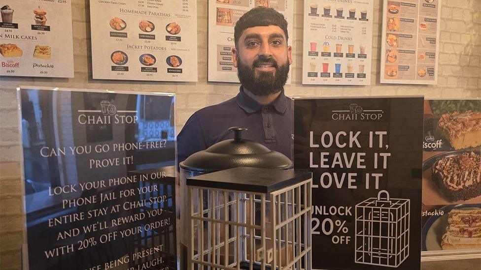 Usman Hussain, who has short dark hair and a beard and is wearing a navy polo shirt, stands in a cafe behind the "phone jail" - a white wire box between two signs that say "lock it, leave it, love it"