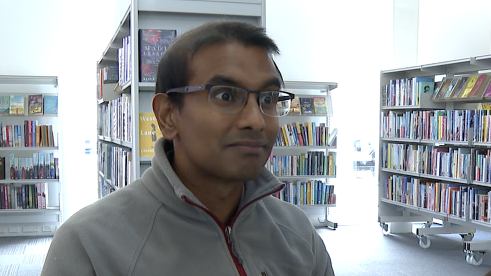 Doctor Kamlesh Sreekissoon is sat in a library with shelves filled with books behind him. He is wearing a fleece and glasses. 
