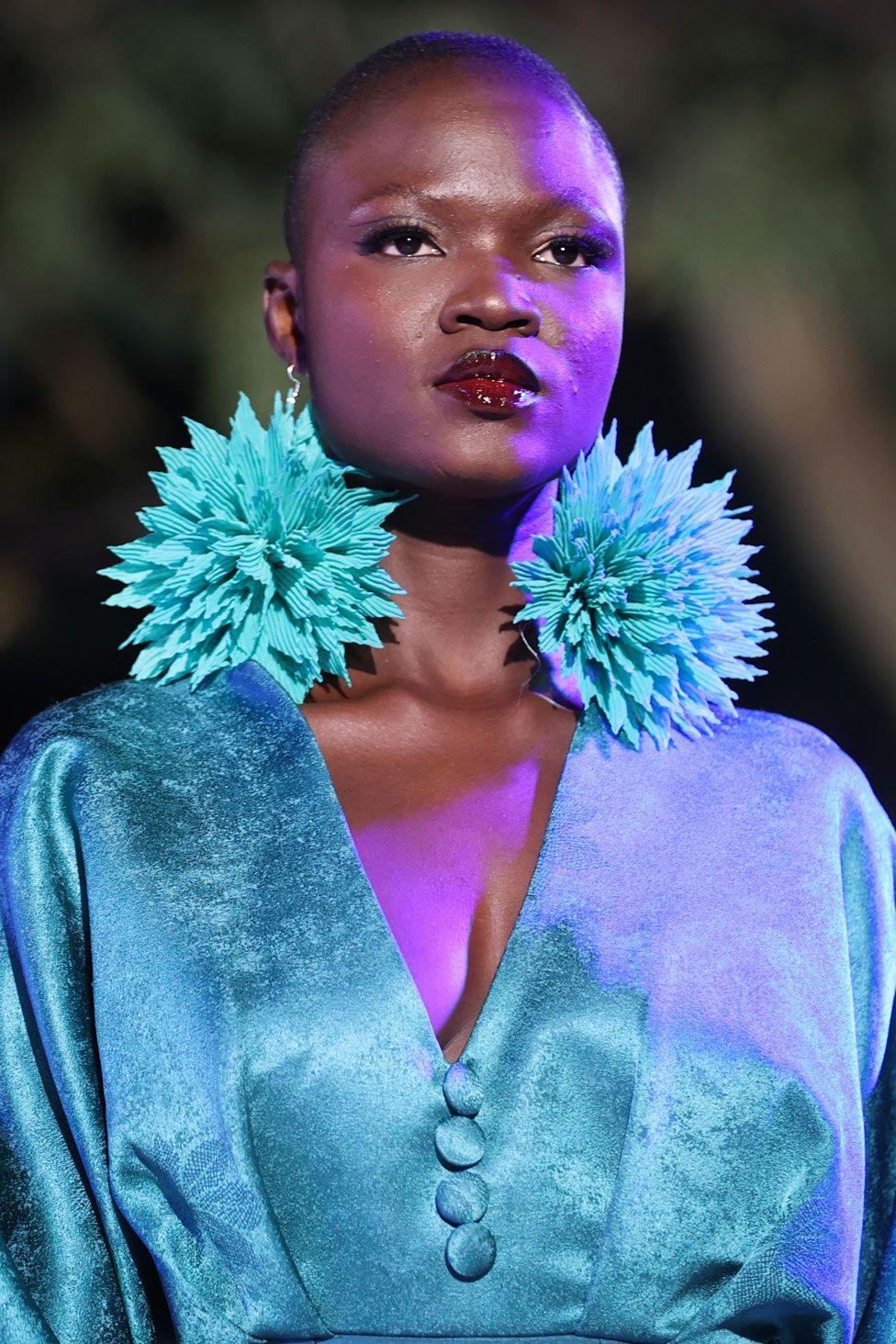 A woman wears a large turquoise pair of pom-pom earrings and a satin dress of a similar shade. Her lipstick is a deep berry colour. In Dakar, Senegal - Saturday 7 December 2024