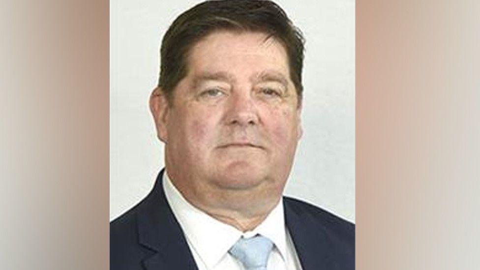 An image of County Councillor Simon Evans, he is not smiling, has dark hair and is wearing a navy suit, white shirt and blue tie.