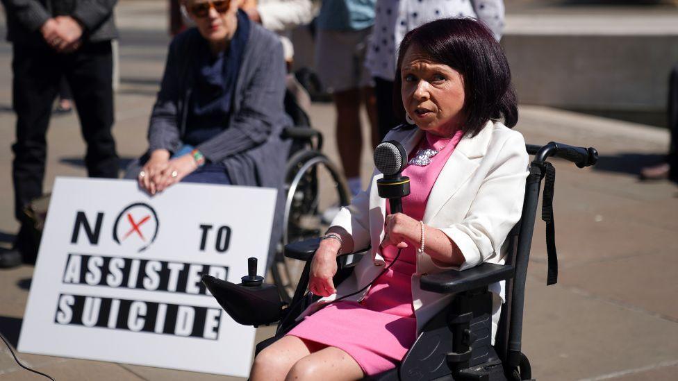 A woman with dark hair, wearing a pink dress and white jacket, speaks into a microphone outside the Scottish Parliament. She is sitting in a black wheelchair. A person behind her is holding a "no to assisted suicide" sign.