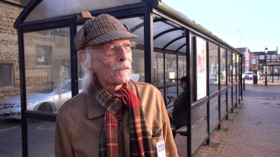 Michael Pollard looking to the right of the camera, with white hair and beard. Behind him is a glass bus stop, where one person sits. 