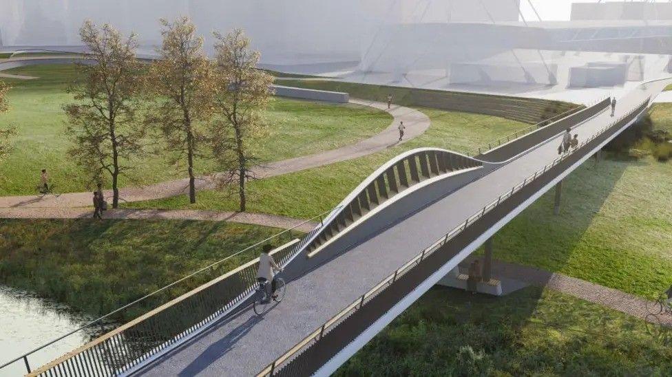 An architect's computed-generated image of a proposed walking and cycling bridge over Grandpont Nature Park in Oxford. The sleek bridge stretches across a field with footpaths, and Oxford Ice Rink can be seen in the background. Trees are also visible near the river which the bridge goes over.