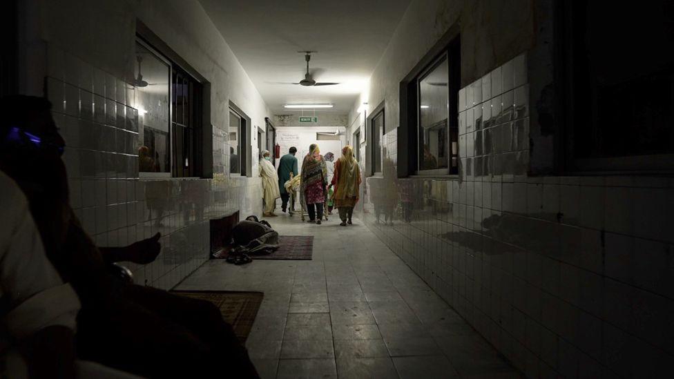 A long, dark hospital corridor with a group of people standing at the far end, and a person lying on a mat on the floor halfway down on the left