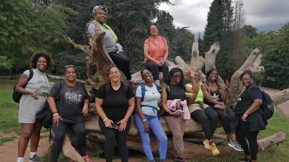 Ten women of colour leaning and sitting on a large tree trunk which has been uprooted out of the ground. They are all wearing casual walking clothes and are smiling at the camera. In the background there are various species of mature trees. 