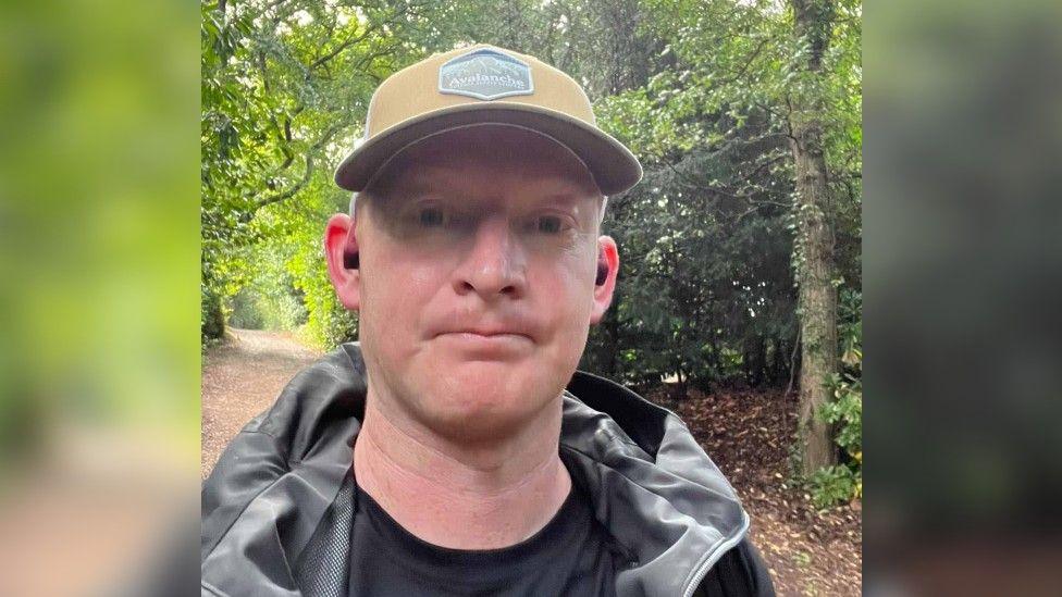 Aaron Oliver looking at the camera is wearing a beige cap and black waterproof jacket, has a woodland in the background.
