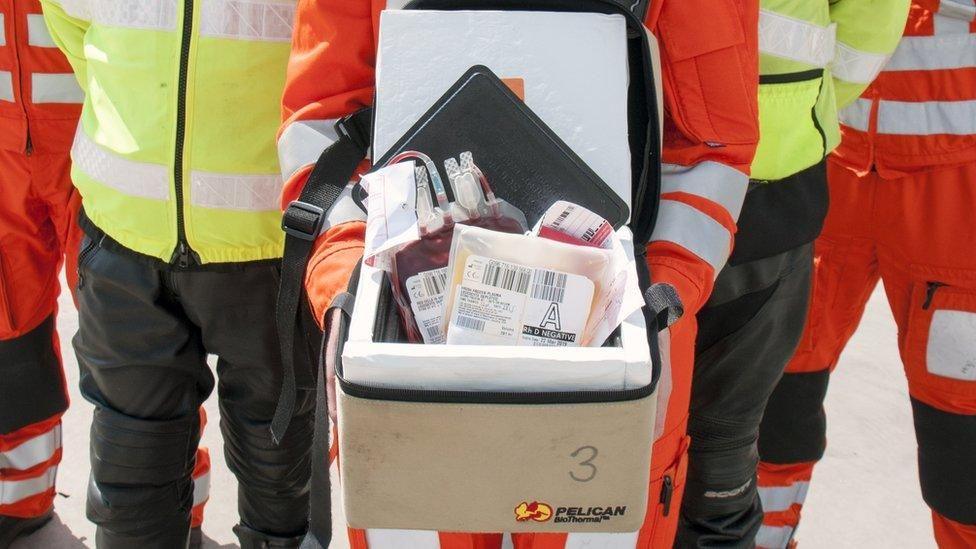 A group of paramedics stand in front of a box of plastic bags containing blood products 