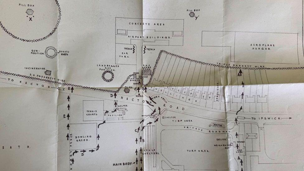 A wartime drawing on cream paper which shows Nacton Road in Ipswich. On the lower level, out of sight, is the main factory for Cranes. On the other side of the road at the top are a row of houses and beyond that the airport. Arrows are drawn across the map showing the routes troops were to take from the factory to the airport. 