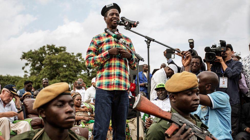 Edgar Lungu speaks at a campaign rally. Dressed in a chequered shirt and beret he is surrounded by photographers and soldiers stand in front of him.
