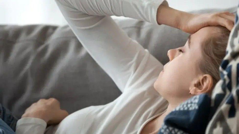 A woman is laying down on a grey sofa. She has her eyes closed and has her hand on her head. She is wearing a white top. 