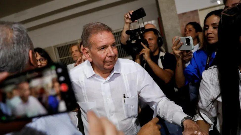 Edmundo González walks past a number of people holding cameras. He has short, brown hair and is wearing a white shirt with a pen sticking out of his front pocket.