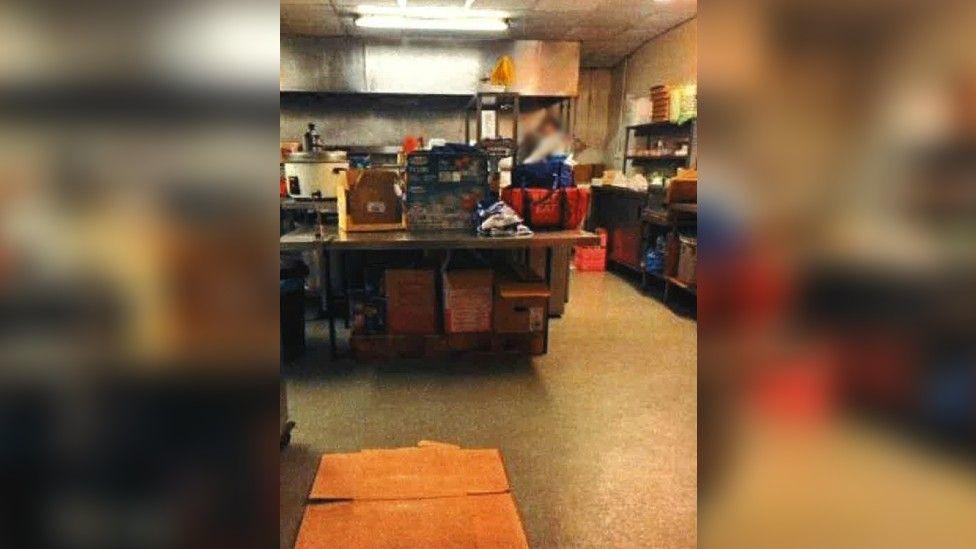 An overcrowded kitchen with boxes on every surface. In the background, the wall is stained black.