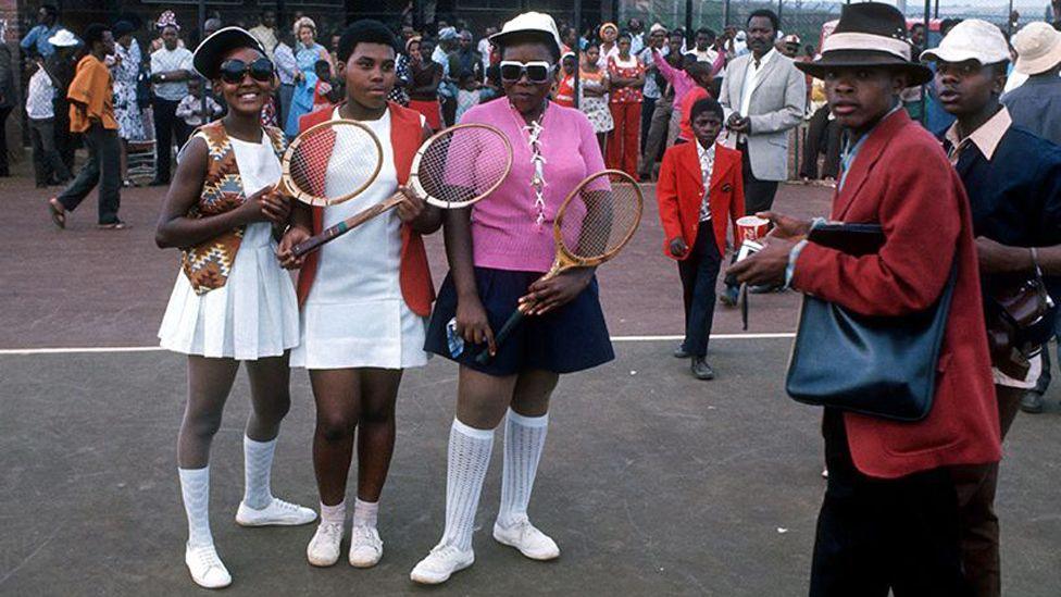 Young girls, some in in tennis whites and sunglasses, pose with racquets as boys in suits and hats walk by. They are part of crowd gathered in Soweto to see Arthur Ashe in November 1973.
