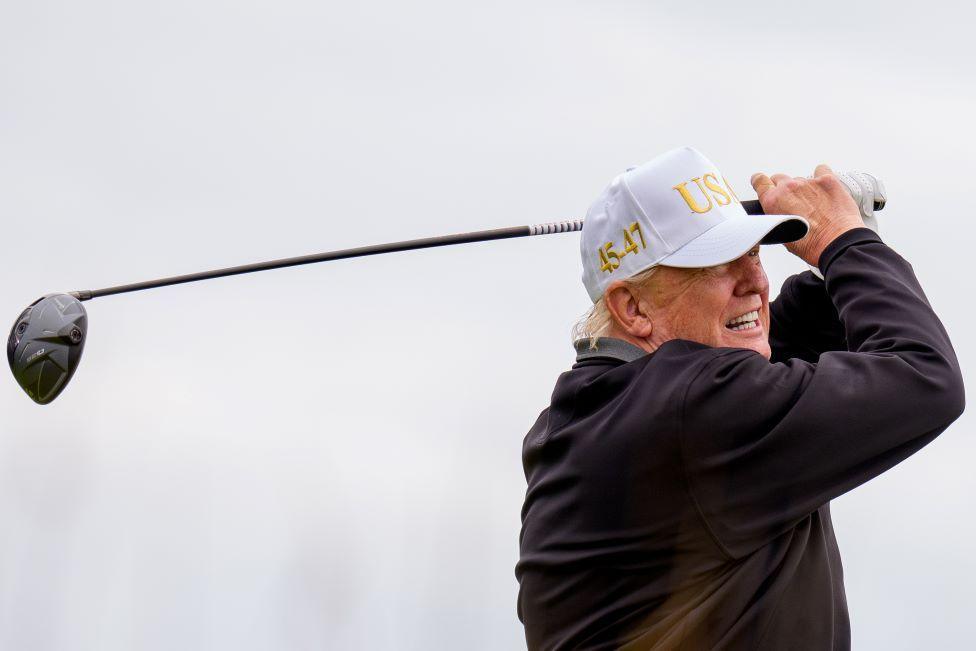 President Donald Trump tees off at a new 18-hole course at Trump International Golf Links in Balmedie, near Aberdeen. He is wearing a white USA baseball cap and is at the end of his golf swing with the driver held aloft behind his head