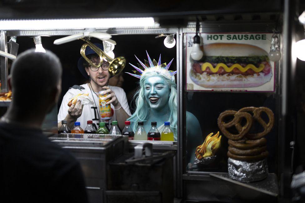 A person dressed as the Statue of Liberty pays for a hot dog at a hot dog cart. A man in glasses and a white t-shirt, holding a hot dog, stands to her right. 