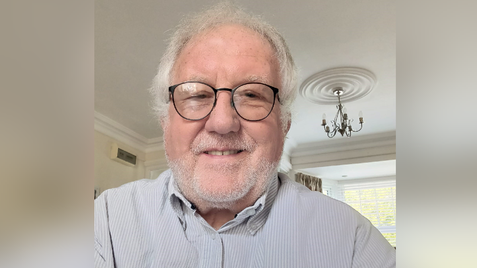 A man with a grey, stubbly beard and white hair looks at the camera. He is wearing black-framed glasses and a white striped shirt.