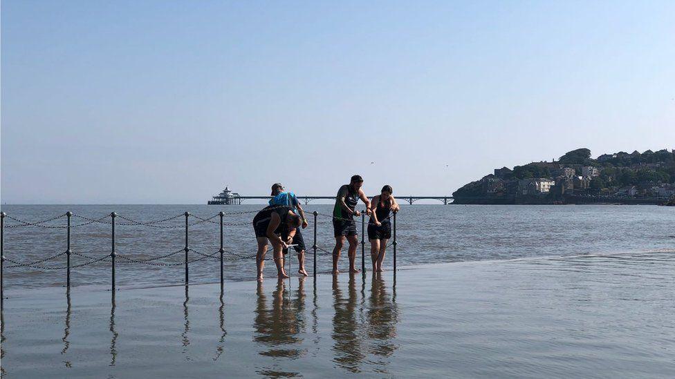 Four people standing near the railings at Clevedon marine lake. The sea and Clevedon Pier can be seen in the background.