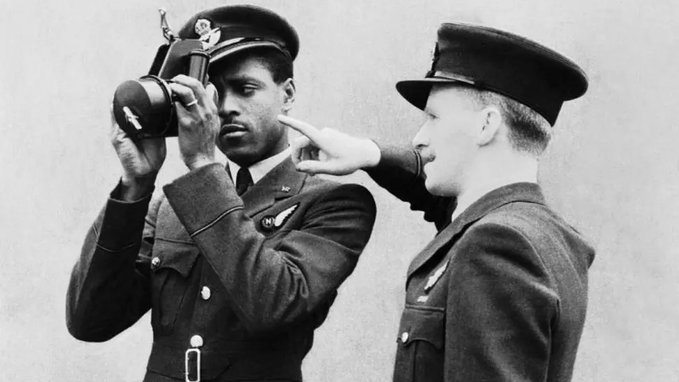 Johnny Smythe stands to the left, he is wearing his RAF uniform of a peaked cap and blazer with a shirt and tie holding a sextant to his right eye and looking through it. Another RAF officer is stood to his right, looking away from the camera and pointing at something out of vision.