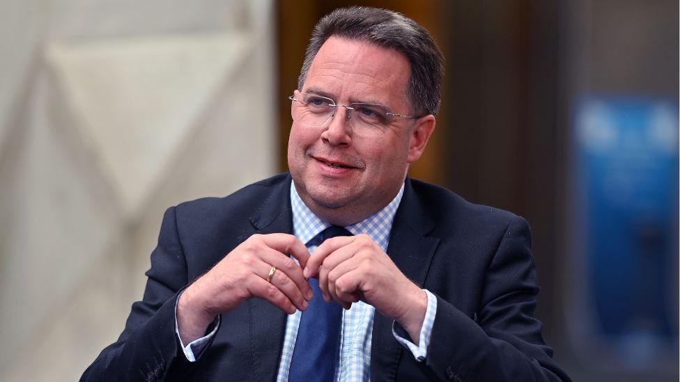 Craig Hoy, a man with dark hair and glasses, sits in the Scottish Parliament. He is visible from the chest up. His hands are touching in front of his chest. He is wearing a dark suit, blue and white chequered shirt and navy blue tie. 