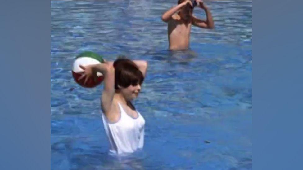 Maggie aged 17 appearing in the film The Givers. She is standing up in an outdoor swimming pool holding a beach ball behind her head, about to throw it. She wears a white vest with blue edging and has dark brown hair in a 1960s bob. She is looking to the right of the shot. In the background also in the pool is a man/boy shading his eyes and wearing no top.