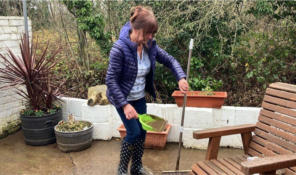 Denise Smith, wearing polka dot patterned wellies, jeans and a blue coat, stands in the garden of her home carrying a muddy dustpan and brush. 