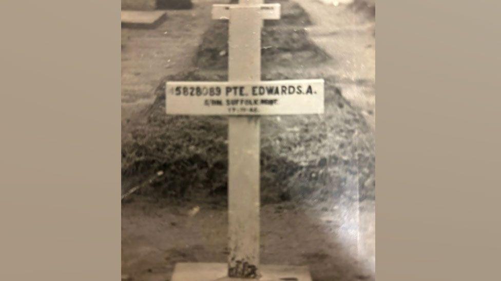A yellowing black and white image of a grave marker. It shows a simple cross, set into the ground. On it says Pte Edwards A, 5th Btn Suffolk Regt and includes his service number. Behind it is some shrubbery and another grave.