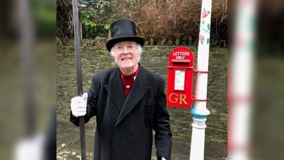 Reg Ling stood next to Frome's Valentine's lamp. He is wearing a long black formal jacket and a black top hat and white gloves