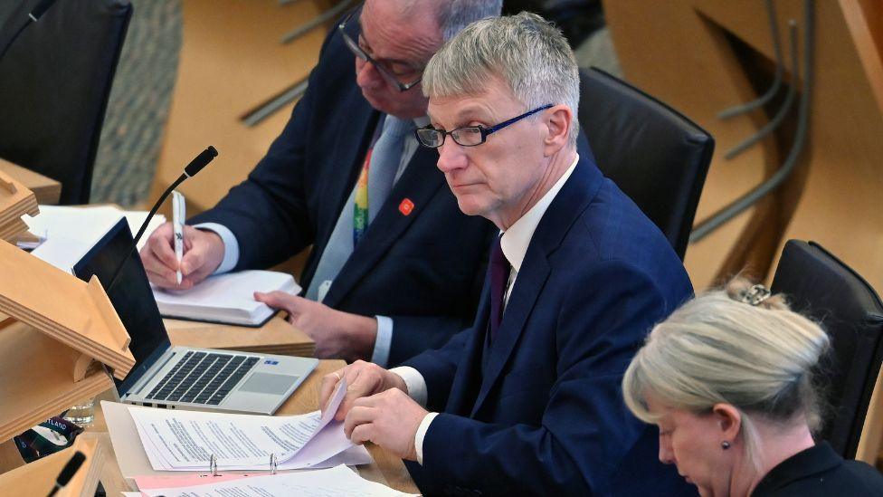 Ivan McKee, a grey haired man with dark-rimmed glasses, leafs through a binder of documents in the Scottish Parliament chamber. He is seated between to other MSPs and is wearing a navy suit, white shirt and purple tie. He is side on to the camera. 