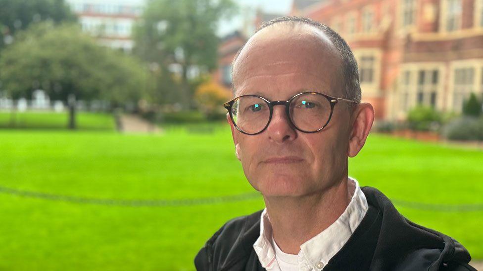Prof Geraint Ellis standing in front of a lawn. He is unsmiling, wearing a white shirt and a hooded top and has horn-rimmed glasses