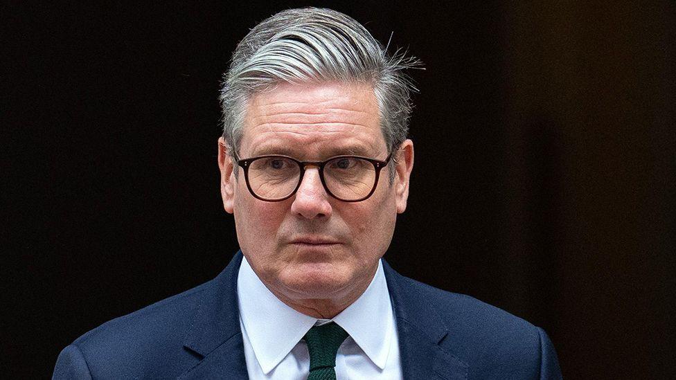 Sir Keir Starmer looks to the left of the camera while wearing suit, tie and glasses as he comes out of 10 Downing Street in London