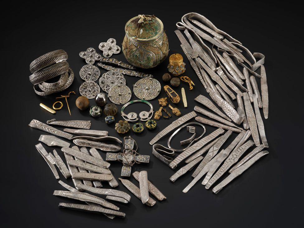 An array of items discovered in a Viking-age hoard in Galloway is spread out on a dark background including an urn, arm rings, bracelets, brooches and other items