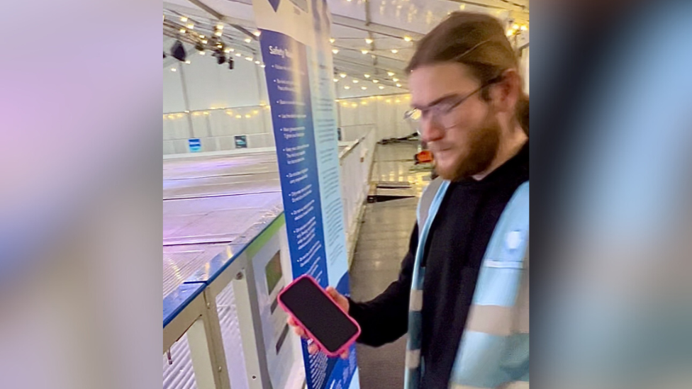 An ice rink worker with long brown hair and glasses holds the pink iPhone next to the rink. He is wearing a black jumper and blue hi-vis jacket.